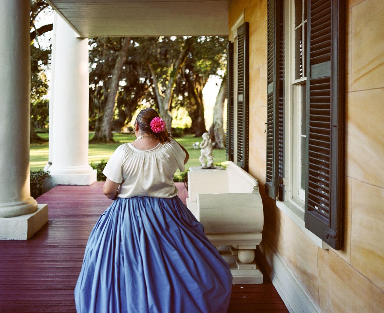 A woman in a long blue skirt and a pink headband stands on the porch of the house. Lafayette, Louisana